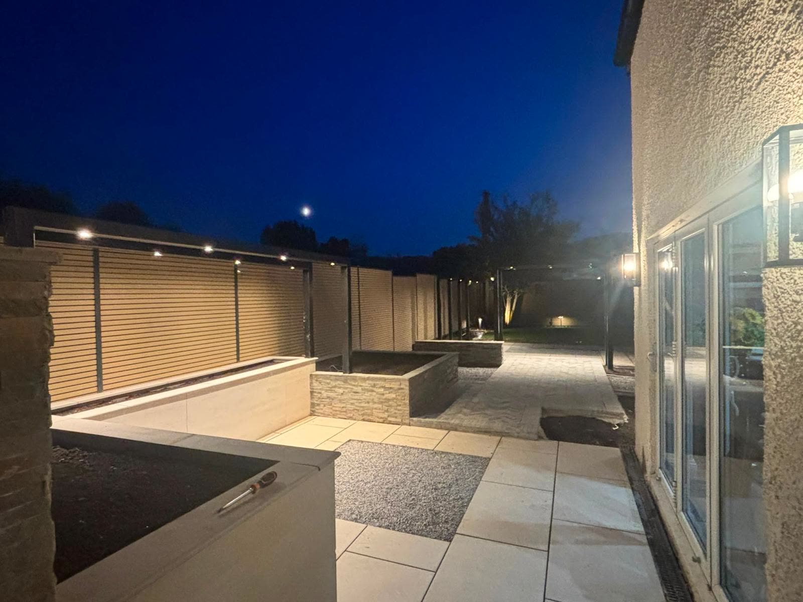 A garden lit at dusk: fence lights, pergola lighting and lit patio